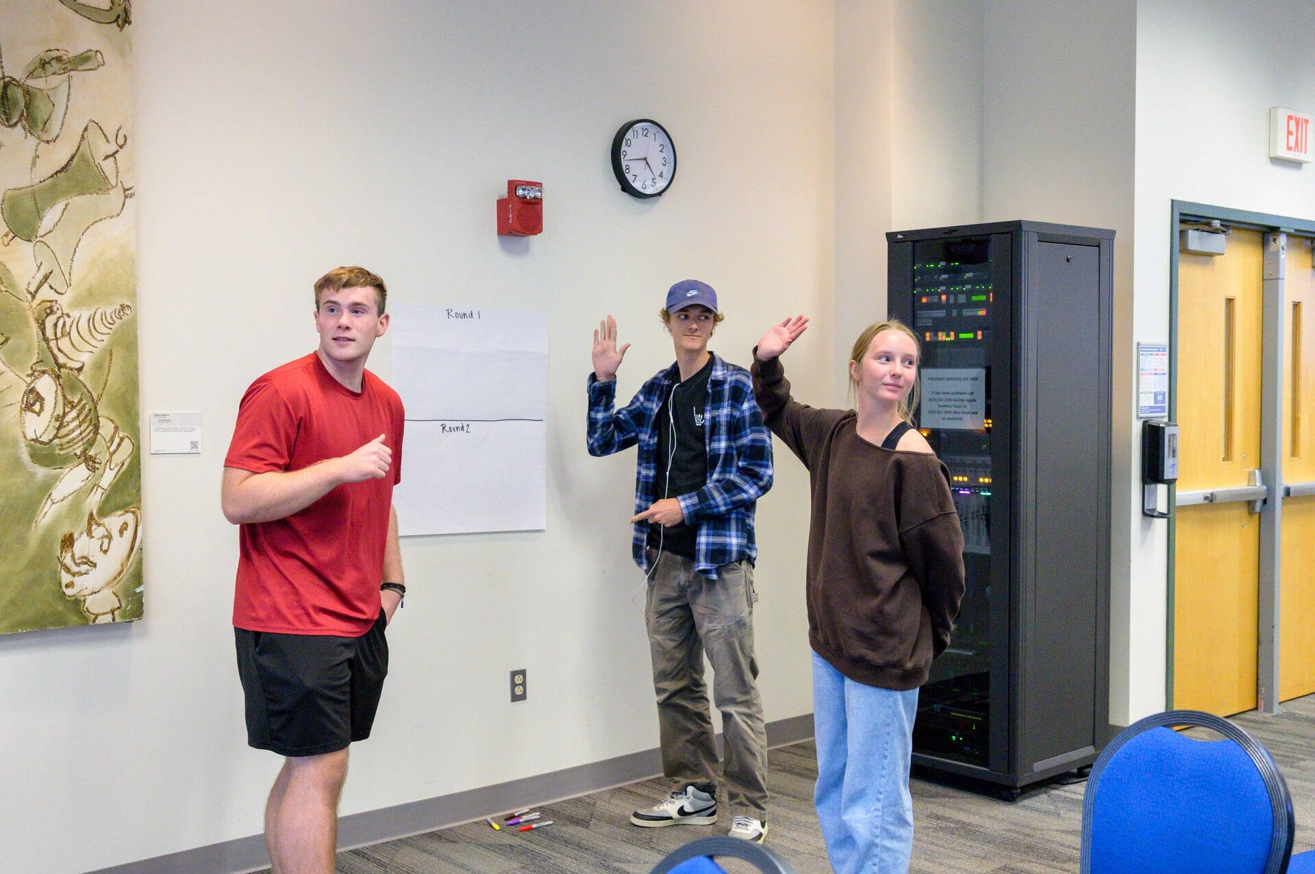 three students stand near a large paper taped to the wall, two on right have hands raised